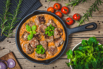 Spicy meatballs with feta cheese in a creamy tomato sauce with herbes on a rustic wooden table, top view