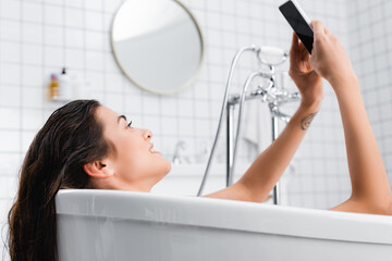 young smiling woman taking selfie while relaxing in bathtub