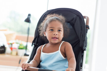 Wall Mural - African American little girl with curly hair sitting and playing on stroller. Cheerful Afro toddler little girl sitting on stroller. Toddler child little girl enjoy, having fun at home