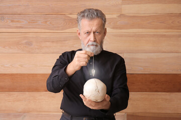 Wall Mural - Male fortune teller with human skull and pendulum on wooden background