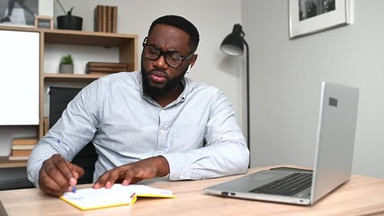 Concentrated guy in glasses is writing some notes in a notebook watching webinar, online lectures on a laptop in. An African-American student studying online