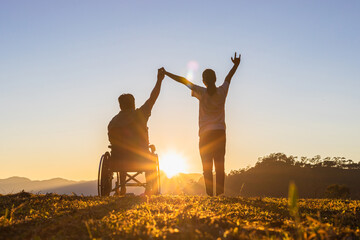 Disabled handicapped young man in wheelchair raised hands with his care helper in sunset.Silhouette