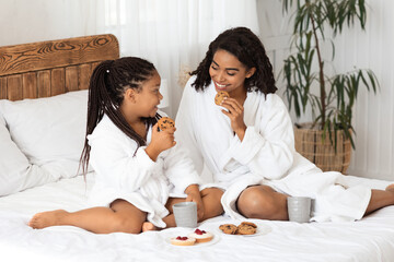 Wall Mural - Cheerful black mom and daughter in bathrobes having tasty snacks in bed