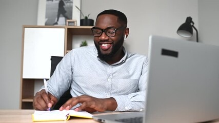 Satisfied African-American young man is taking a notes watching online webinars, classes, coach lectures. Smiling multiracial millennial using laptop sitting at the desk and writing in a notebook