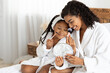 © Prostock-studio - African mother and her little daughter in bathrobes making makeup on bed