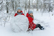 Two twin girls in red jackets rolling huge snowball to make snowman in a snowy forest