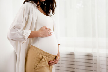 Portrait shot of young beautiful woman on second trimester of pregnancy. Close up of pregnant female in casual attire with arms on her round belly. Expecting a child concept. Background, copy space.