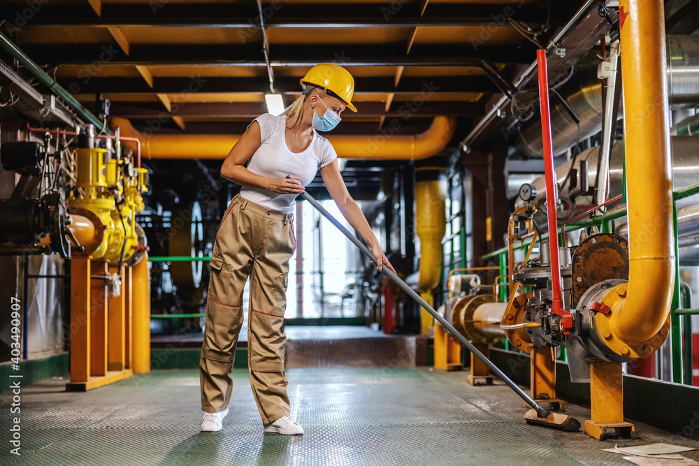 Tidy hardworking female worker in working suit with protective helmet on head and face mask brooms heating plant facility during corona outbreak. - obrazy, fototapety, plakaty 