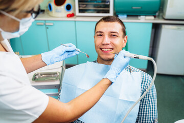 A young, attractive European-looking guy came to the dentist. Sits in a dental chair. The dentist bent over him and prepares to treat problem teeth. Happy patient and dentist concept