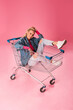 Beautiful fashionable young blonde woman sitting on a shopping trolley in a studio. Wearing jeans and pink top. Spring, summer fashion photo over pink background.