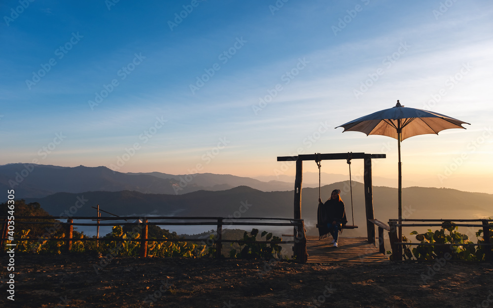 A female traveler sitting on a swing while watching a beautiful view and sunrise in the morning - obrazy, fototapety, plakaty 