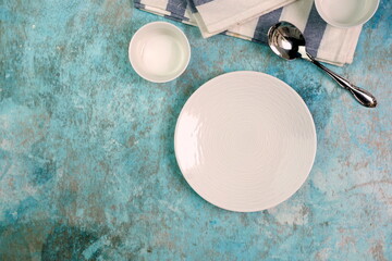 White empty ceramic plates on grainy texture blue background with stripes napkin, bowl and silver spoon as serving food preparation concept. Top view of white tableware 