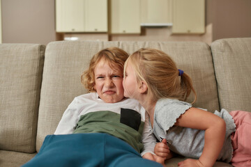 Portrait of little boy looking disgruntled while his sibling sister kissing him on cheek, cuddling together on a sofa at home