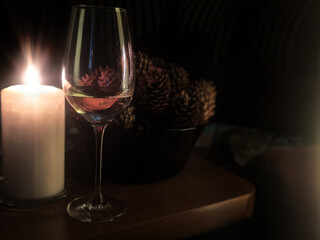 Glass of white wine on wooden table near glowing candle and bowl with heap of pine cones behind on dark background