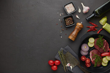 Beef steaks and cutting vegetables on grill pan, olive oil and spices on black slate background. Top view. Steak menu