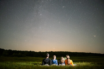 Parents and their kids resting in night field observing dark sky with many bright stars.