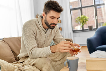 health, cold and people concept - sick young man in blanket drinking hot tea with lemon and honey at home