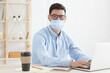 © Damir Khabirov - Young employee sitting in office, working with laptop, wearing mask, practicing social distancing because covid safety protocol