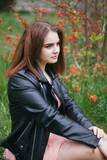 Fototapeta Młodzieżowe - Portrait of a young girl. The girl is sitting on a background of red flowers.A young girl in a leather jacket.