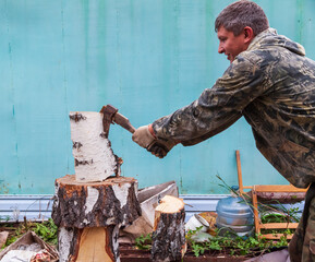 A log split in half with an ax on a deck with male hands in cloth gloves and a camouflage jacket in the backyard. Household work to prepare for the winter.