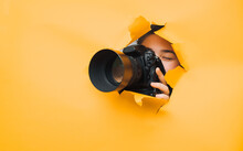 A Teenage Girl Is Holding A Camera With A Telephoto Lens That Looks Out Through A Torn Hole In Yellow Paper. Concept Of Photo Reporter, Paparazzi, Espionage, Yellow Press.