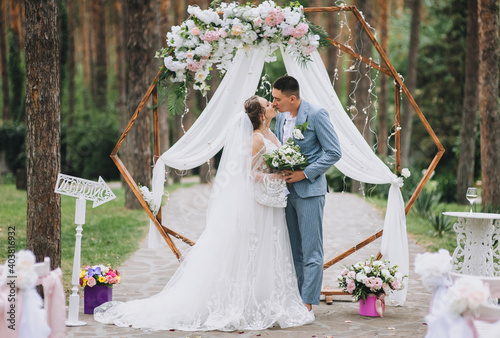 Stylish groom in a blue striped jacket hugs and kisses the bride in a lacy white dress near a wooden arch decorated with flowers at the wedding ceremony. Beautiful portrait of newlyweds.