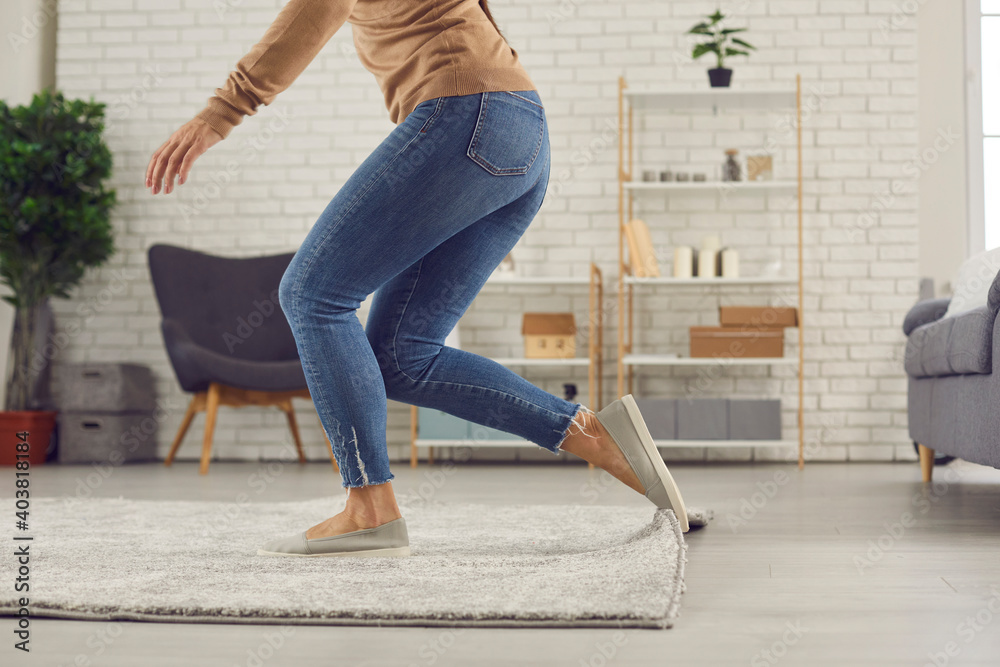 Unrecognizable female trips over rug. Clumsy faceless woman in uncomfortable shoes stumbles on rug in living-room and is about to fall on floor. Domestic accidents and getting injured at home concept - obrazy, fototapety, plakaty 