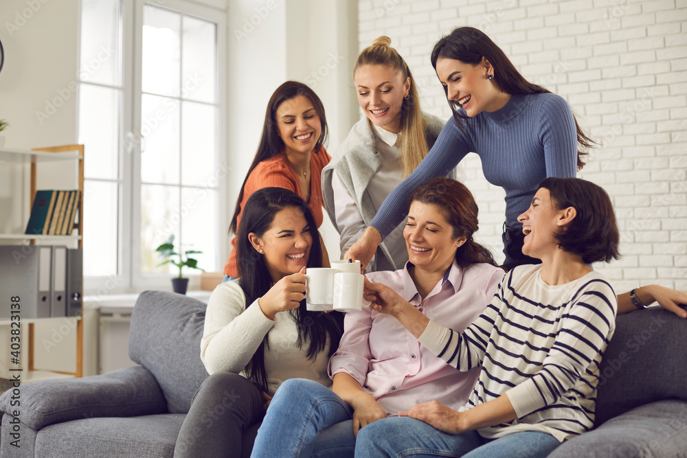 Group of happy young women having fun and enjoying time together. Smiling friends or colleagues sitting on sofa at home or in office, laughing at funny joke, clinking cups and drinking coffee or tea - obrazy, fototapety, plakaty 