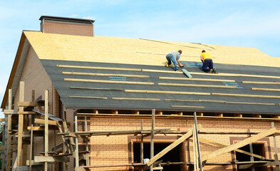 A close-up of a brick house under construction with the roofing contractors on unfinished rooftop installing roofing waterproofing rubberized asphalt underlayment before asphalt shingles installation.