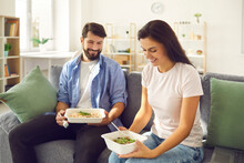 Couple Eats Fresh Takeaway Food From Plastic Containers They Ordered From The Delivery Service. Happy Man And Woman Enjoying Dinner Sitting At Home On Sofa. Eating And People Concept.