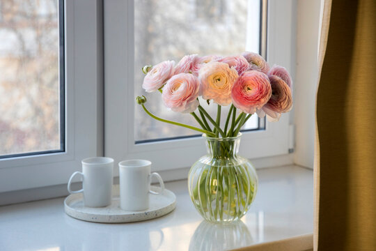 Pink Persian buttercups on a white artificial stone window sill behind a curtain. Two white cups with tea are on the round tray