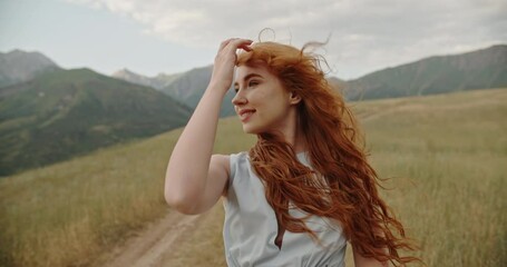 Poster - Giel with red hair walking down the wheel trails on meadow with scenic mountain background, looking at camera and smiling - freedom, tranquility concept 4k footage