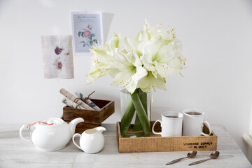 Wall Mural - A bouquet of white lily in a glass vase on a table with two tall cups of coffee, a teapot, spoons, and a milk jug. Copy space