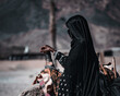 Unidentified bedouin woman in an abaya standing near a lying camel with her hand on a camel saddle