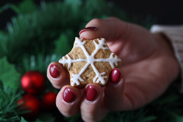 Woman hand holding a cookie in the shape of a snowflake and green Christmas tree in the background. Holiday sweet food concept