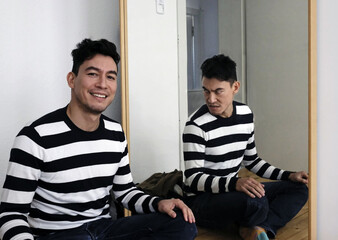 Caucasian smiling man sitting in front of a mirror while his alter-ego frowns at him in the mirror