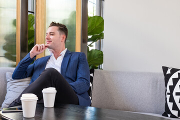 Adult Caucasian businessman dress in smart formal suit sitting inside modern office space drinking coffee while waiting and planning for business meeting. Entrepreneur concept with copy space.