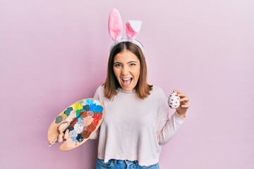 Sticker - Young beautiful woman wearing cute easter bunny ears holding painting palette and egg celebrating crazy and amazed for success with open eyes screaming excited.