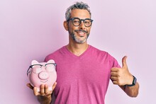 Middle Age Grey-haired Man Holding Piggy Bank With Glasses Smiling Happy And Positive, Thumb Up Doing Excellent And Approval Sign