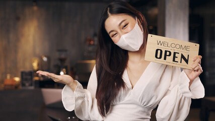 Business owner attractive young Asian woman in apron hanging we're open sign on front door  welcoming clients to new cafe. Happy waitess with protective face mask holding open sign at cafe .