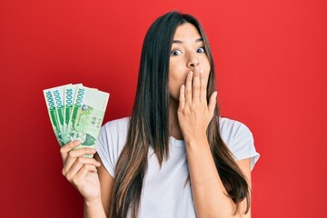 Wall Mural - Young brunette woman holding 1000 south korean won banknotes covering mouth with hand, shocked and afraid for mistake. surprised expression