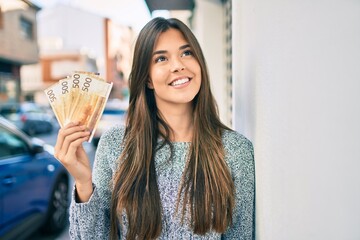 Wall Mural - Young beautiful hispanic girl smiling happy holding norwegian krone banknotes at the city.