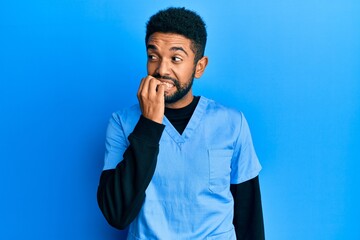 Wall Mural - Handsome hispanic man with beard wearing blue male nurse uniform looking stressed and nervous with hands on mouth biting nails. anxiety problem.