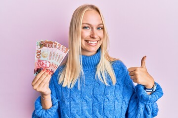 Sticker - Young blonde girl holding 100 new zealand dollars banknote smiling happy and positive, thumb up doing excellent and approval sign