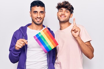 Poster - Young gay couple holding rainbow lgbtq flag smiling with an idea or question pointing finger with happy face, number one