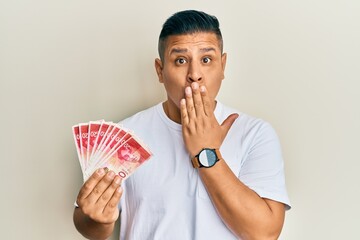 Canvas Print - Young latin man holding 20 israel shekels banknotes covering mouth with hand, shocked and afraid for mistake. surprised expression