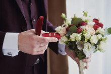 Man's Hands Holding A Red Wedding Rings Box And A Wedding Bouquet. Preparations For The Wedding.