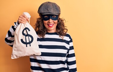 Middle age brunette burglar woman wearing mask and cap holding bag with dollar symbol looking positive and happy standing and smiling with a confident smile showing teeth