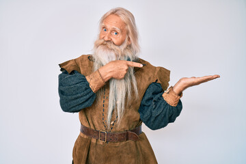 Old senior man with grey hair and long beard wearing viking traditional costume amazed and smiling to the camera while presenting with hand and pointing with finger.