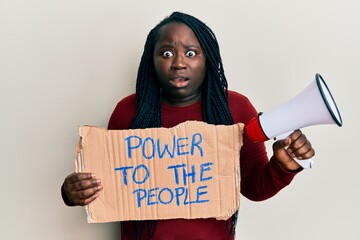 Sticker - Young black woman with braids holding power to the people banner and megaphone afraid and shocked with surprise and amazed expression, fear and excited face.